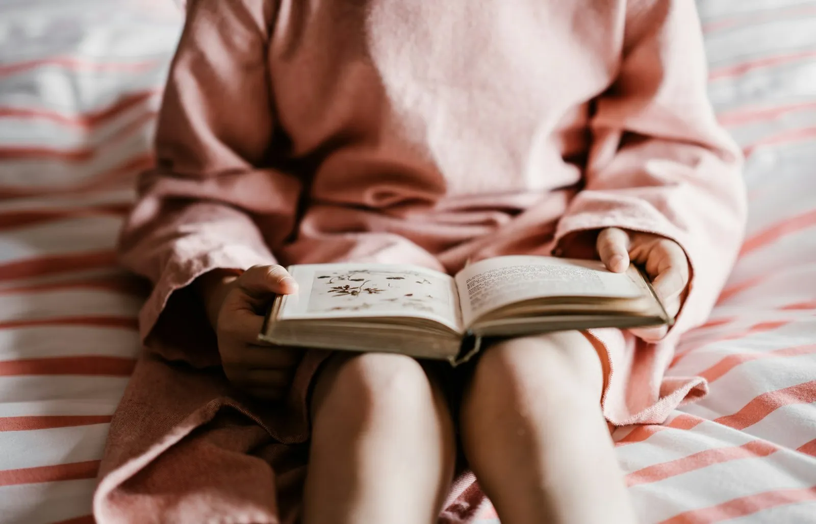 A child in pink pyjamas sitting on a red and white striped bedspread, holding open a picture book on their knees.