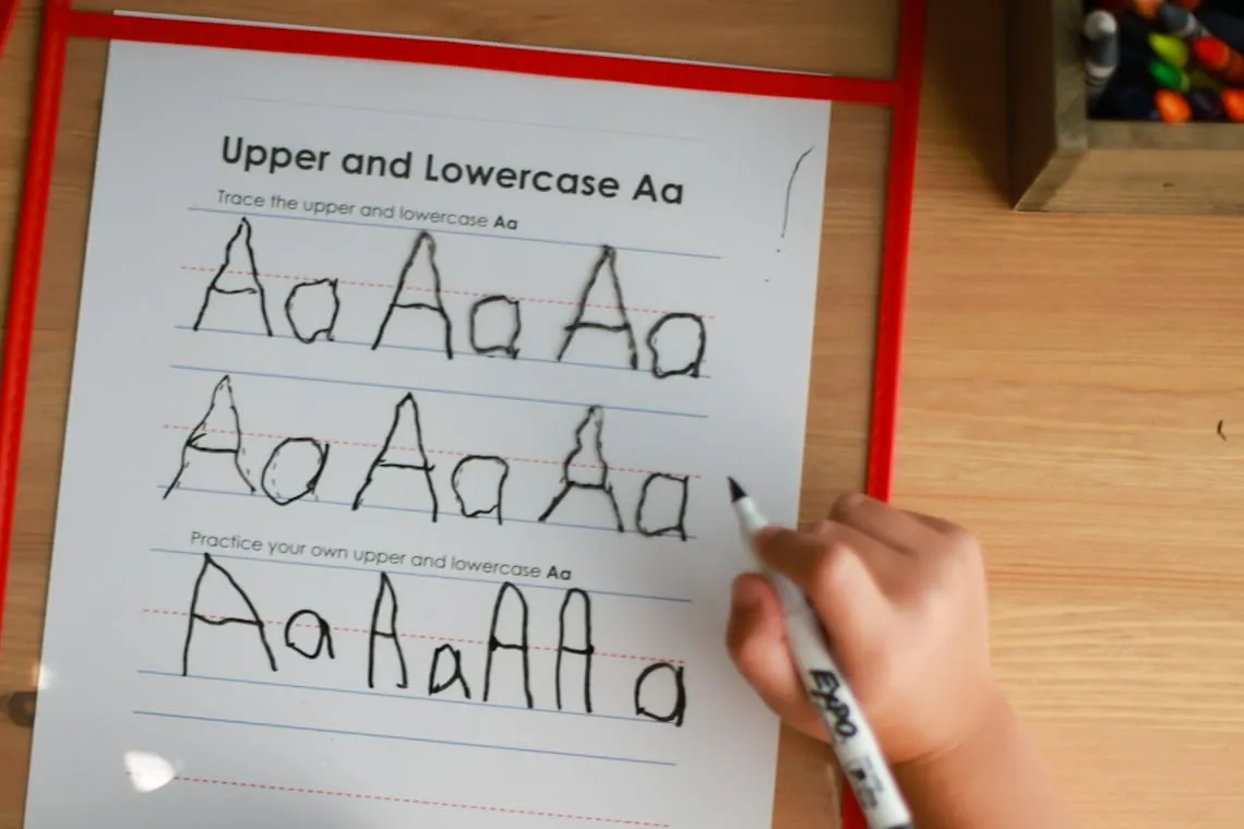 A child writing capital and lowercase A on a ruled practice sheet with a dashed midline: two rows traced over printed models, then a row of their own letters underneath, noticeably less even than the traced ones.