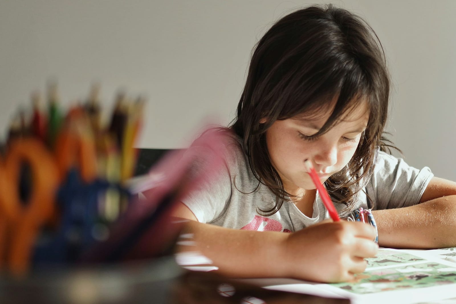 A young girl leaning over a workbook at a table, writing with a red pencil, concentrating hard, a pot of colored pencils out of focus behind her.