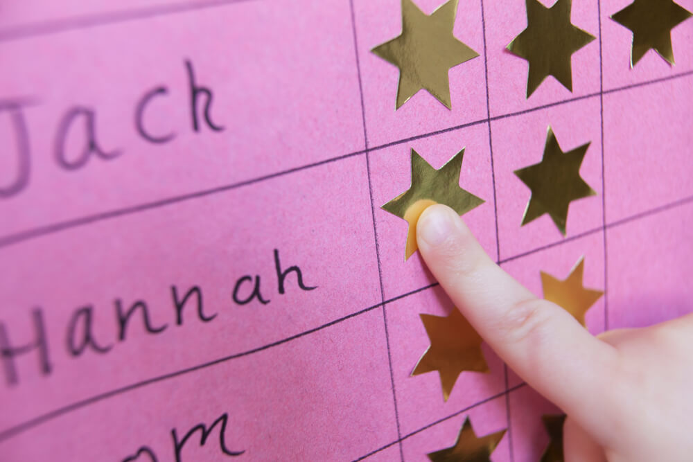 Close-up of a child's finger pressing a gold star sticker onto a pink reward chart with handwritten names.